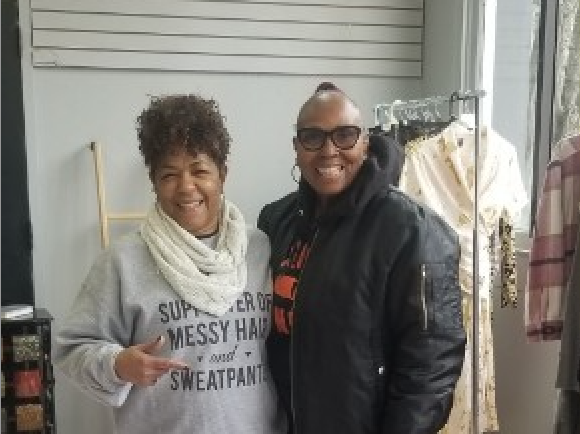 Community in action: two smiling women in the boutique; one in a grey sweatshirt and white scarf, one in a black bomber jacket and glasses