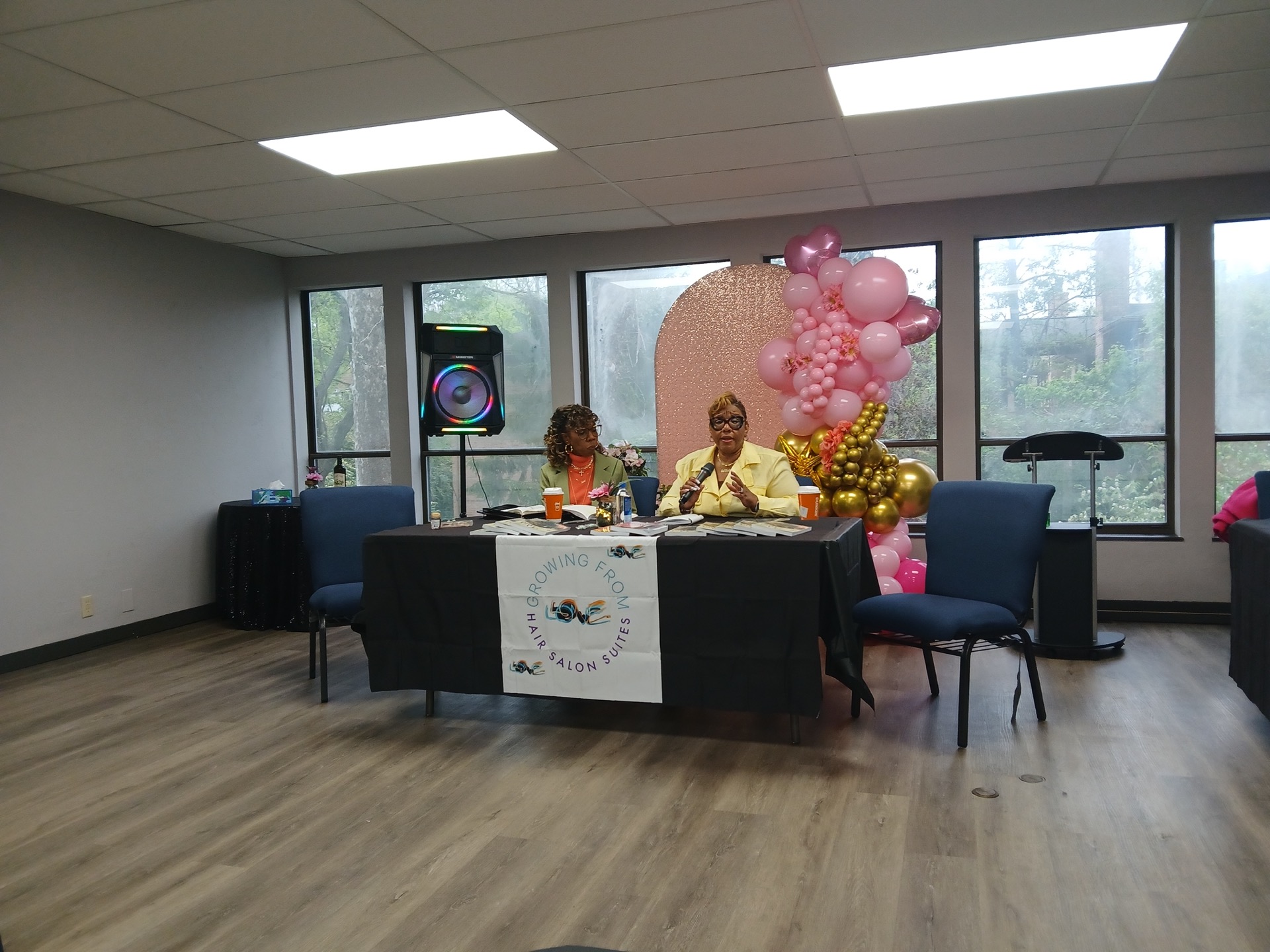 Growing From Love Hair Salon Suites event: three panelists at a table with banner, pink backdrop, and balloon decor