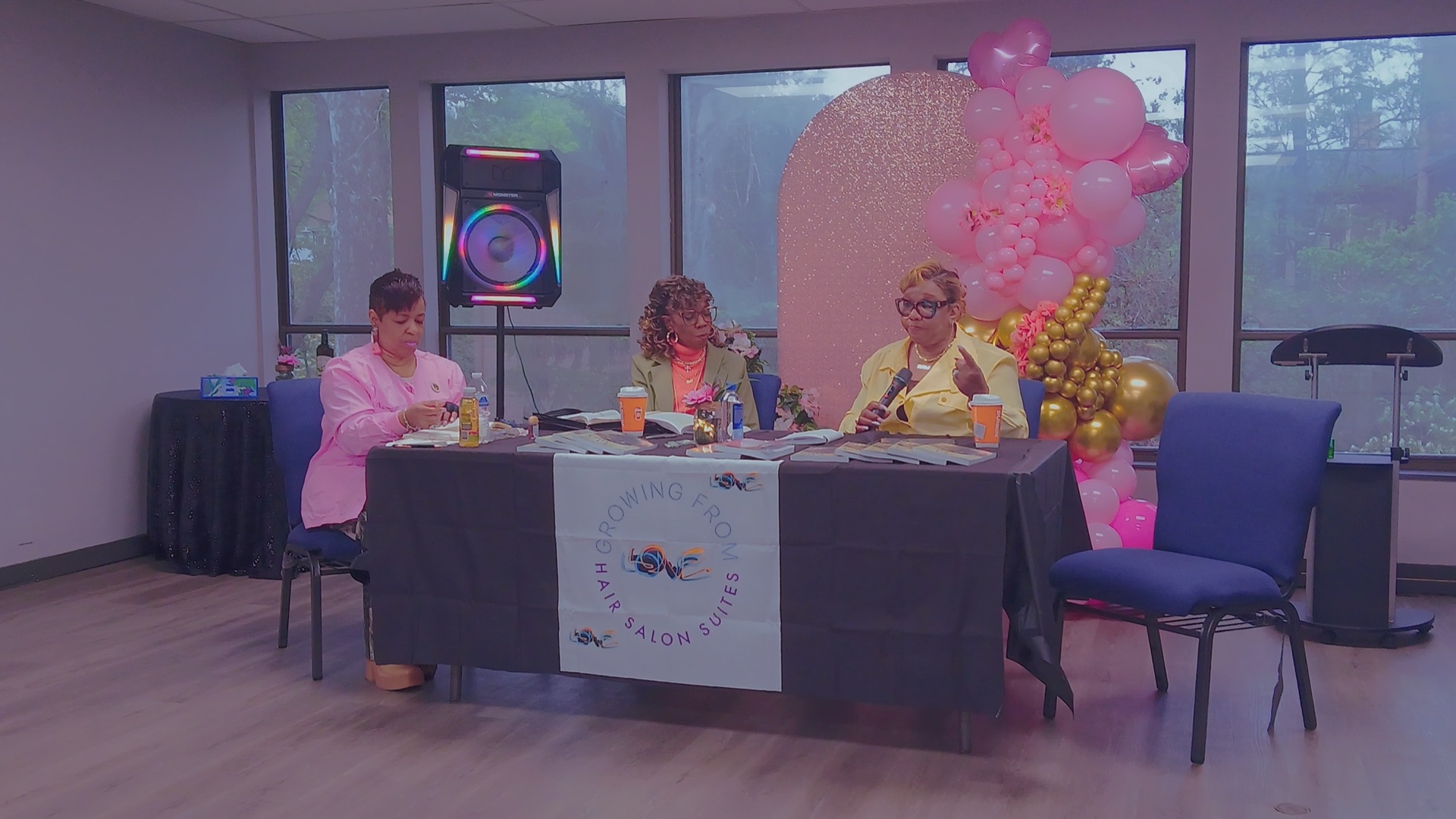 Growing From Love Hair Salon Suites gathering: speakers at table with LOVE logo banner and festive room setup